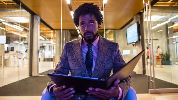 LaKeith Stanfield sits in an office holding an open folder