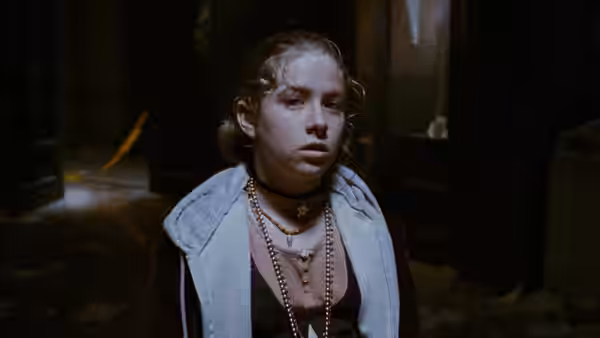 Sarah (Rochelle Davis) stands in a dim room wearing layered necklaces