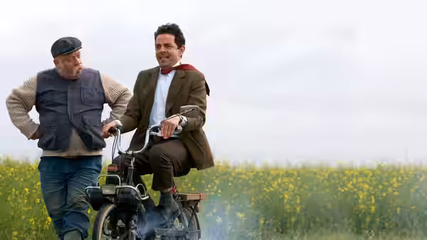 Mr. Bean (Rowan Atkinson) rides a small motorcycle through a field beside a local man