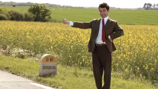 Mr. Bean (Rowan Atkinson) points along a roadside beside a field of yellow flowers