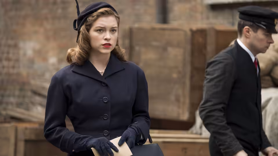 Young Joan (Sophie Cookson) holds papers beside a uniformed man on a city street