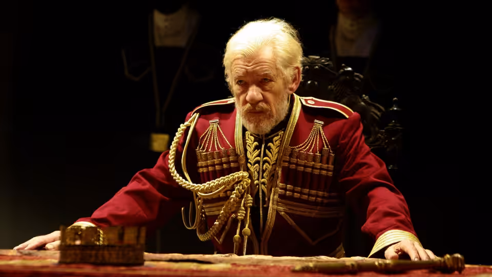 King Lear (Ian McKellen) leans over a table in an ornate red military uniform