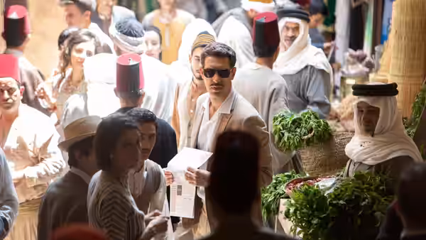 Eli Cohen (Sacha Baron Cohen), wearing sunglasses, carries papers through a crowded market