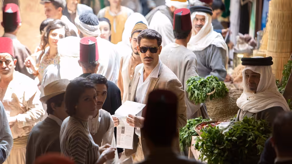 Eli Cohen (Sacha Baron Cohen), wearing sunglasses, carries papers through a crowded market