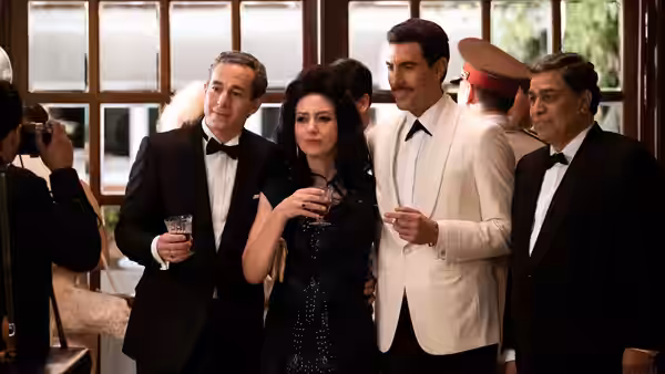 A mustached man in a white suit poses with formally dressed guests holding drinks
