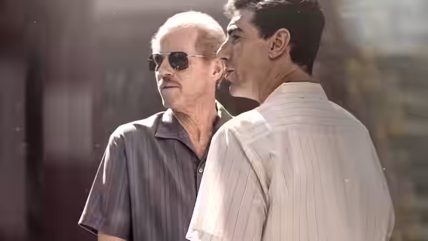 Two men stand close together outdoors, one wearing sunglasses and a dark shirt