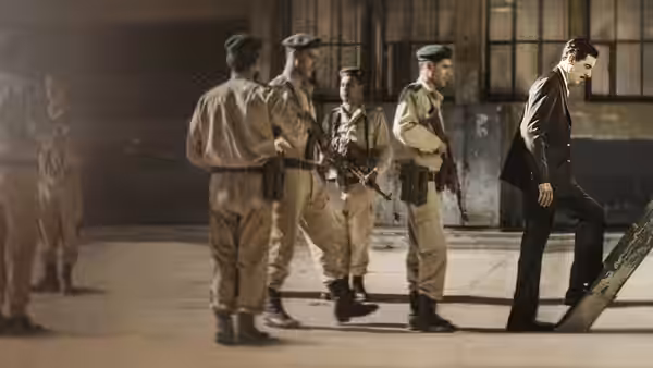 Eli Cohen (Sacha Baron Cohen) walks past uniformed soldiers in a guarded compound