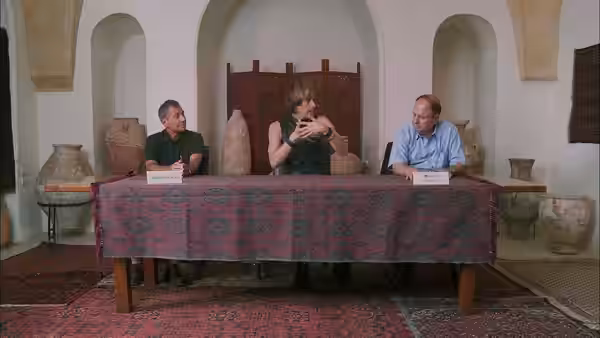 Brüno (Sacha Baron Cohen) gestures between two men seated at a table
