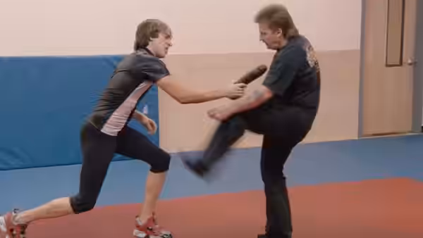 Brüno (Sacha Baron Cohen) holds a microphone as a man kicks toward him