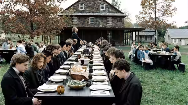 Villagers gather at long outdoor tables for a meal beside a stone house