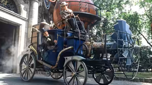 Two men ride an elaborate steam-powered carriage along a tree-lined street
