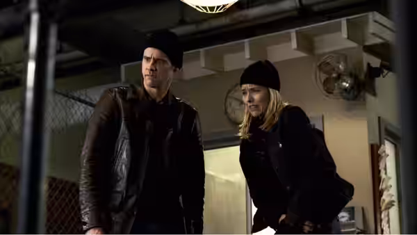 Dick and Jane Harper stand in black clothes inside a dim industrial room