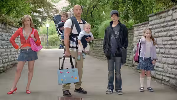 Shane Wolfe (Vin Diesel) carries a baby and a diaper bag among children on a tree-lined path