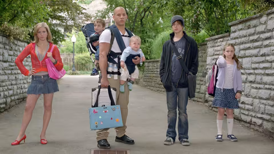 Shane Wolfe (Vin Diesel) carries a baby and a diaper bag among children on a tree-lined path