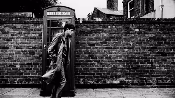 man in long coat stands beside a telephone booth on pavement