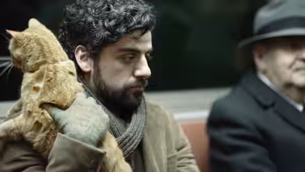 Llewyn Davis (Oscar Isaac) holding an orange cat on public transport beside an older man