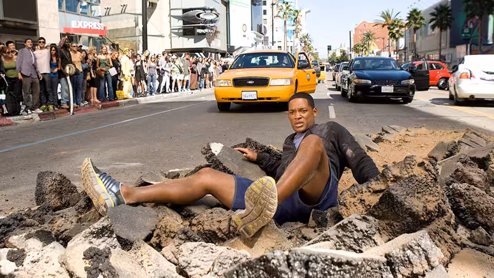John Hancock (Will Smith) sits amid broken pavement as traffic and onlookers gather behind him