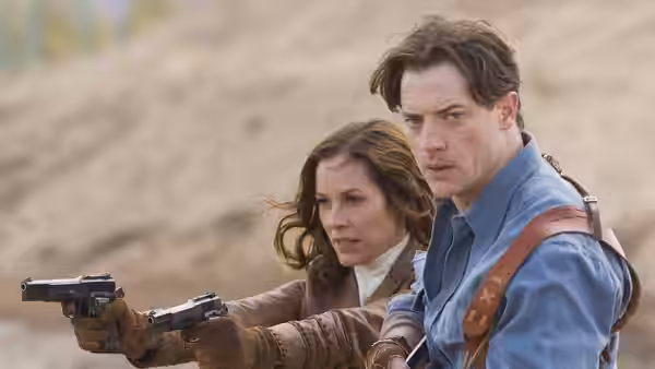 Rick and Evelyn O'Connell aim pistols outdoors against a sandy hillside