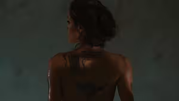 A tattooed woman stands with her back turned in a dim room