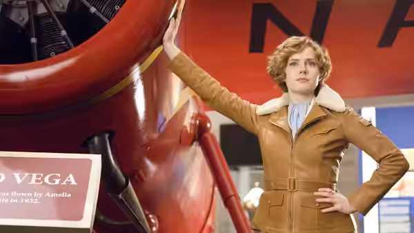 Amelia Earhart poses beside a red airplane with one hand on its wing