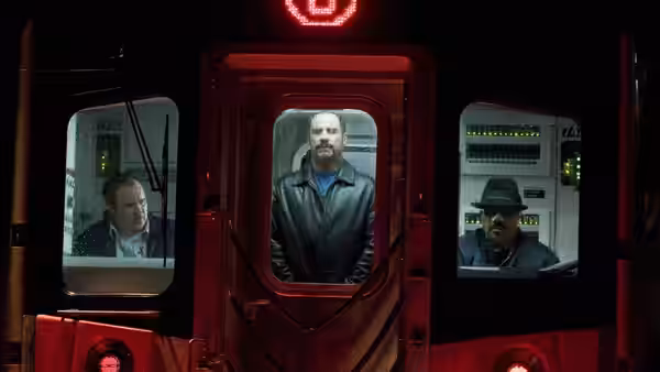 Ryder (John Travolta) stands in a subway car doorway between two seated men
