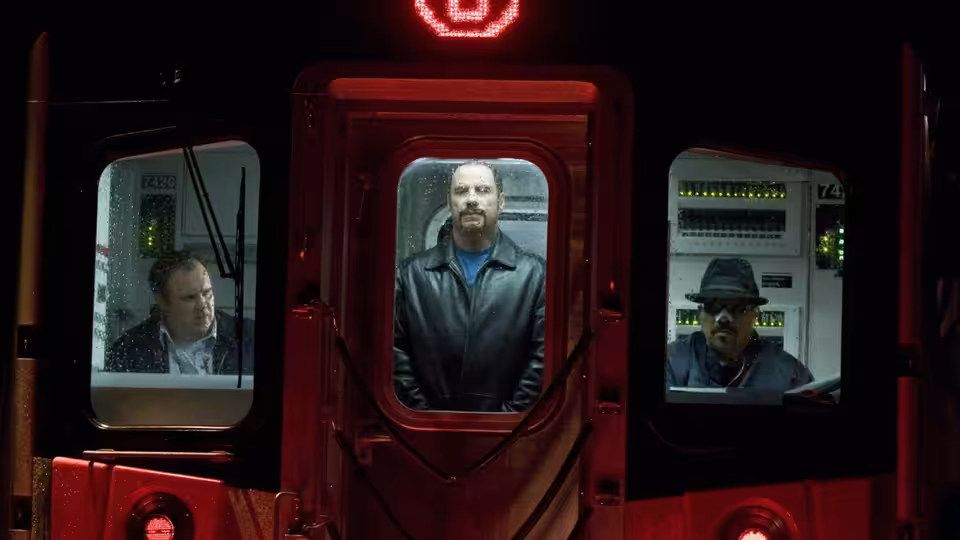 Ryder (John Travolta) stands in a subway car doorway between two seated men