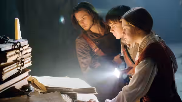 Caspian, Edmund and Lucy gather around open books and a candle in a dim room