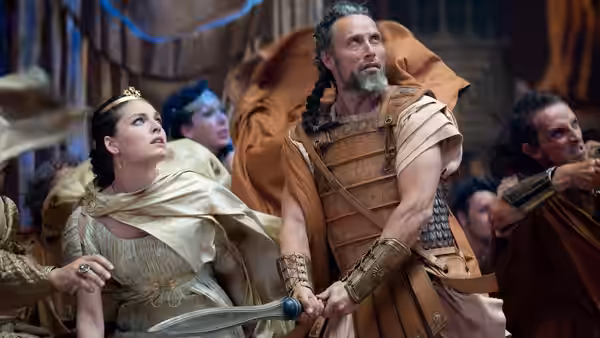 A warrior stands beside a crowned woman as they look upward in a crowded hall