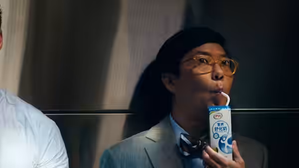 A bespectacled man in a suit sips from a carton inside an elevator