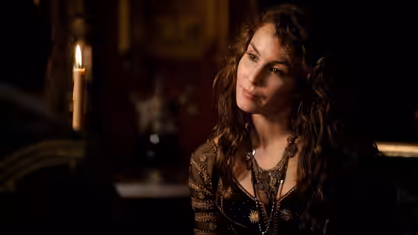 A woman in a dark embroidered dress sits by candlelight and looks toward someone off-frame