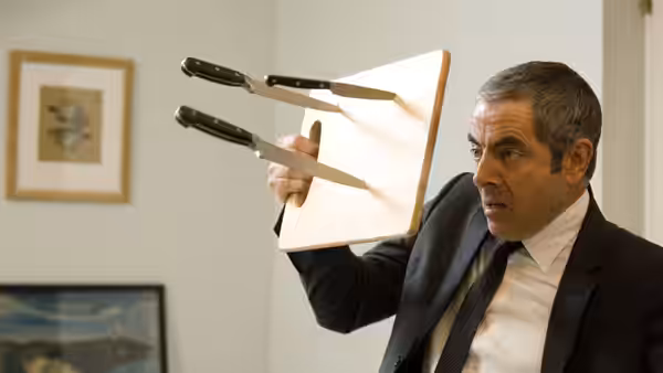Johnny English (Rowan Atkinson) holds up a clipboard to deflect two flying knives