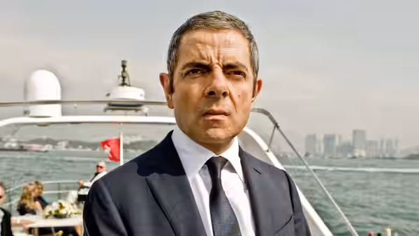 Johnny English (Rowan Atkinson) stands aboard a yacht with the city waterfront behind him