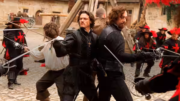 Aramis and Athos fight guards with swords in a courtyard as D’Artagnan joins them