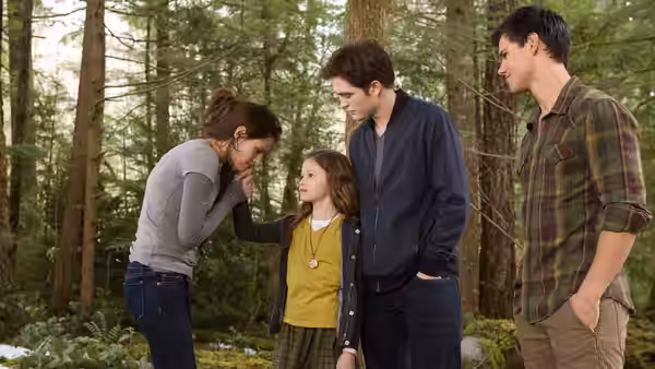 Bella Swan (Kristen Stewart) bends toward her daughter as Edward and Jacob stand nearby in the forest