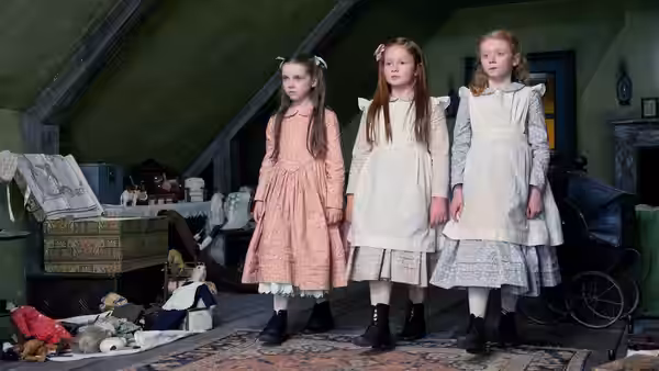 Three girls in pale dresses stand among scattered toys in a cluttered attic