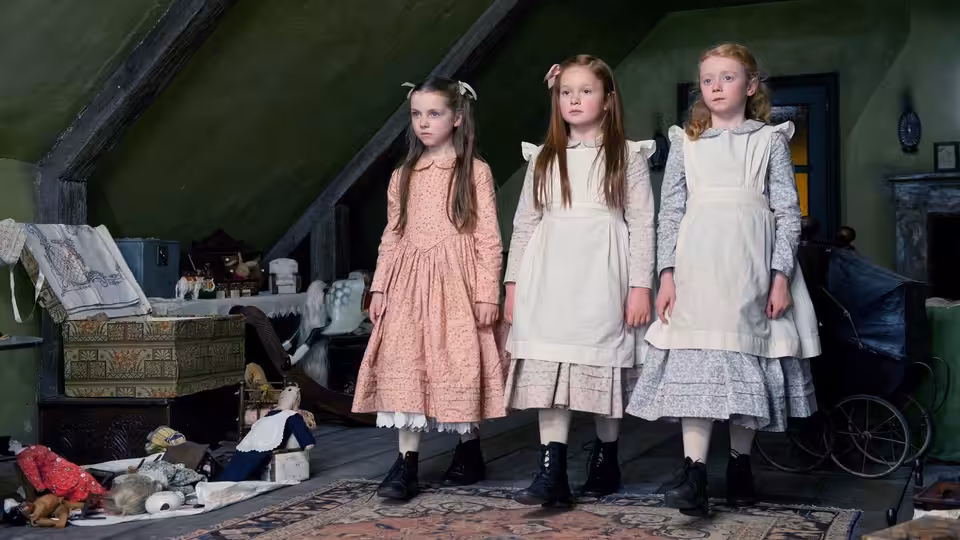 Three girls in pale dresses stand among scattered toys in a cluttered attic