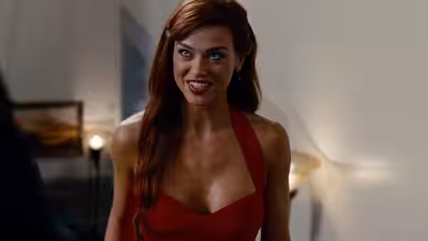 A red-haired woman in a red dress leans forward in a hotel room
