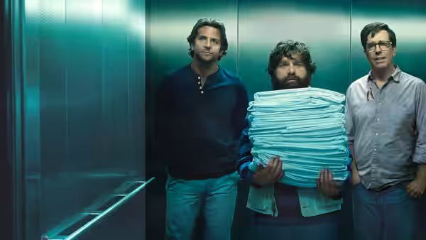 Alan carries a tall stack of folded towels between Phil and Stu in an elevator