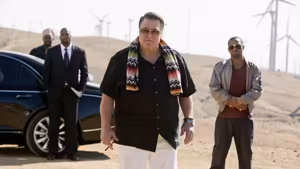 Marshall walks toward the camera with bodyguards beside a parked car in the desert
