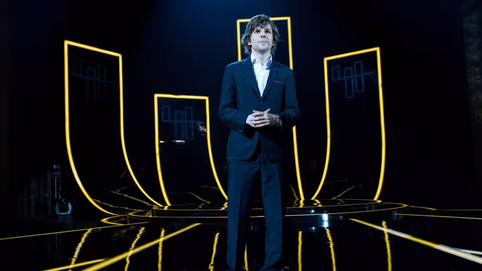 J. Daniel Atlas (Jesse Eisenberg) stands on a stage framed by glowing yellow panels