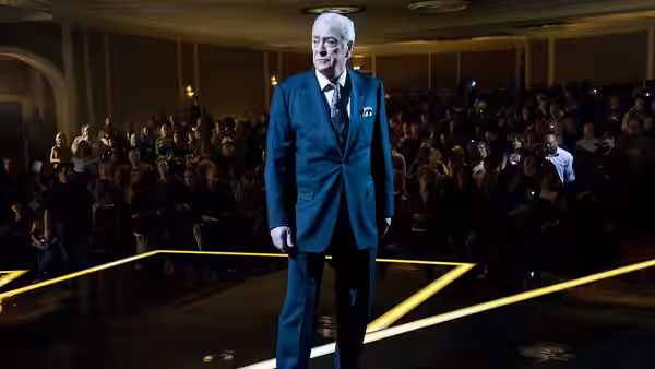 Arthur Tressler (Michael Caine) stands before a seated audience onstage