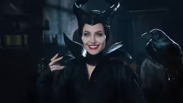 Maleficent (Angelina Jolie) smiles beside Diaval in raven form indoors