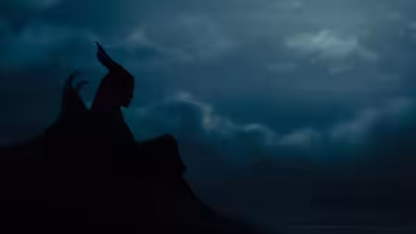 Maleficent sits in silhouette on a rocky ledge beneath a cloudy sky