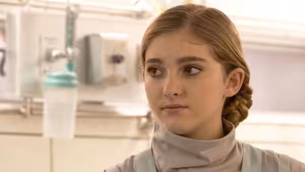 A young woman in a pale uniform looks aside in a bright medical room