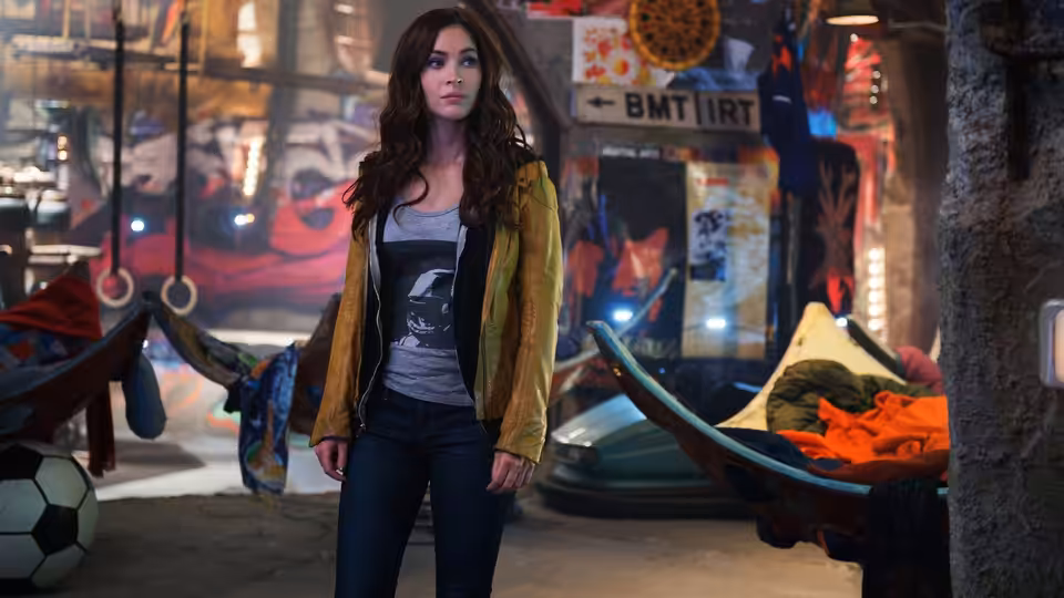 April O'Neil (Megan Fox) stands among hammocks and scattered belongings in the Turtles' underground lair