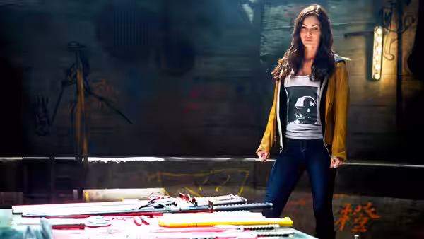 April O'Neil (Megan Fox) stands beside a table displaying weapons