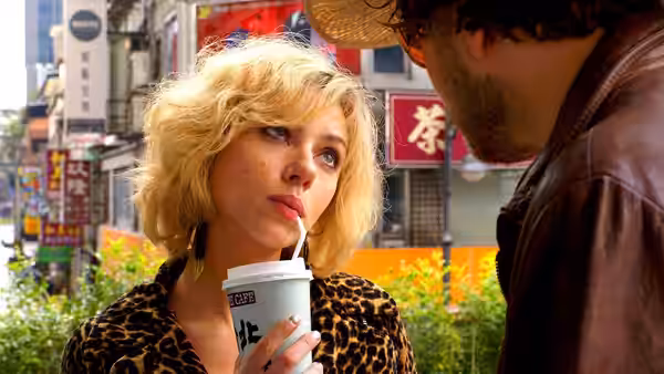 Lucy (Scarlett Johansson) drinks through a straw while talking with a man on a city street