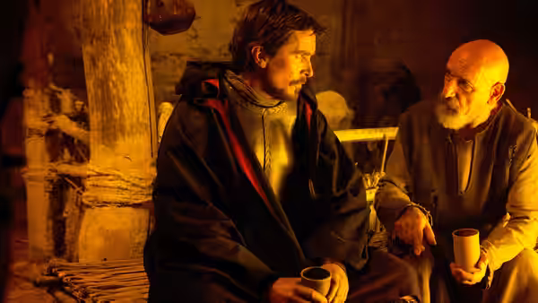 Moses (Christian Bale) sits with an elderly bearded man in a warmly lit stone dwelling