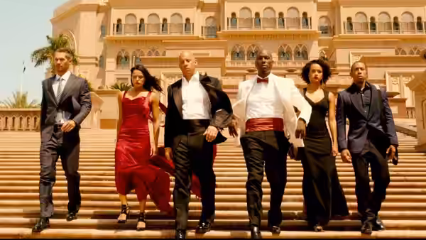 A group of six sharply dressed people descend the steps of an ornate palace