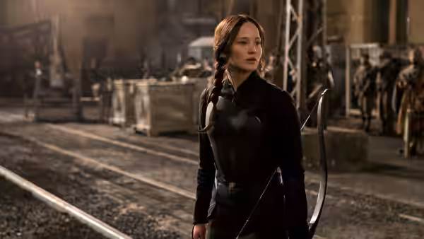 Katniss Everdeen (Jennifer Lawrence) stands beside railway tracks as armed fighters gather behind her